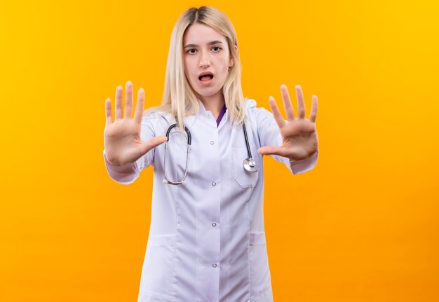 Sad doctor young girl wearing stethoscope in medical gown showing stop gesture with both hands on isolated yellow background