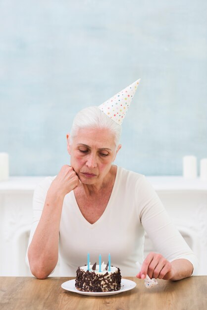 Грустная старшая женщина смотрит на день рождения торт со свечой над столом