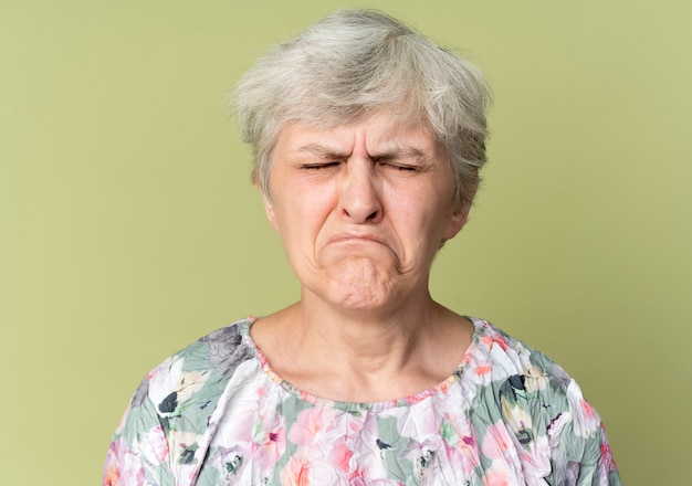 Sad elderly woman stands with closed eyes isolated on olive green wall