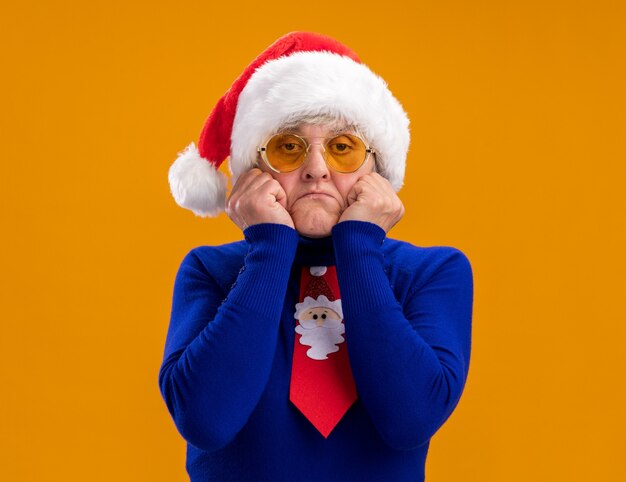 Sad elderly woman in sun glasses with santa hat and santa tie puts hands on face isolated on orange wall with copy space