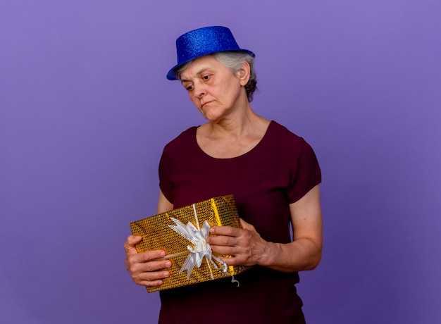 Sad elderly woman wearing party hat holds gift box looking down isolated on purple wall with copy space