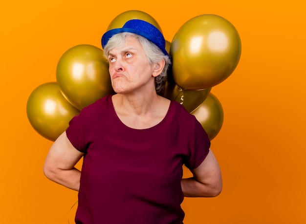 パーティーハットをかぶった悲しい年配の女性は、オレンジ色を見上げて後ろにヘリウム気球を保持します