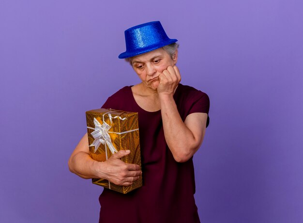 Sad elderly woman wearing party hat puts hand on chin and holds gift box isolated on purple wall with copy space