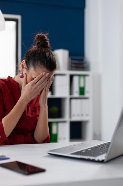 Sad entrepreneur in red shirt holding face in palms. disappointed businesswoman working on laptop recieved bad news. stressed employee having a breakdown sitting at startup desk.