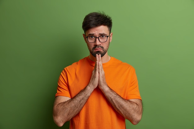 Sad European man with hopeless upset expression prays and has hope for better, guilty look, asks for forgiveness, feels really sorry, presses hands together says please forgive me wears orange t shirt