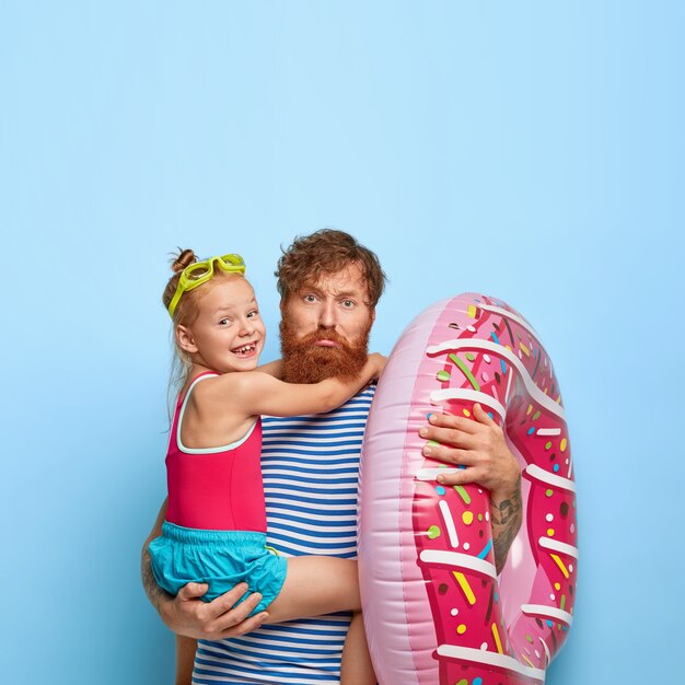 Sad fatigue father with ginger beard, carries small daughter on hands, inflated swimring, going to beach together, dressed casually, entertain in resport place