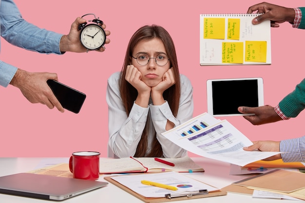 Sad female workaholic keeps hands under chin, busy making project work, studies papers, wears elegant white shirt, sits at desktop, unknown people stretch hands with notes, alarm clock, smartphone