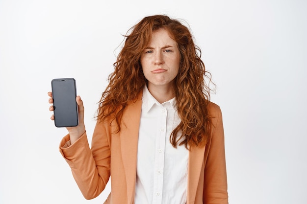 Sad frowning business woman showing mobile phone screen and sulking dislike smartphone app standing in suit against white background