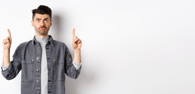 Sad frowning guy pointing and looking up standing hesitant on white background