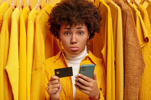 Sad frustrated Afro woman makes payment via online wallet, holds credit card and modern cellphone