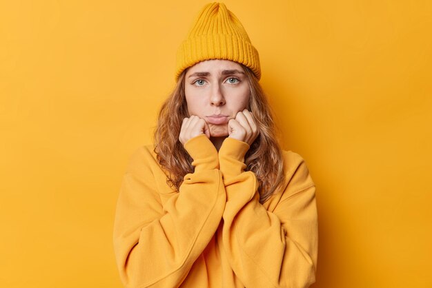 슬픈 좌절한 여성이 턱 아래에 손을 얹고 입술이 노란색 배경에 격리된 모자와 점퍼를 입은 불만스러운 표정을 하고 있습니다. 부정적인 감정과 감정