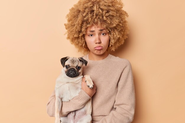 Sad frustrated woman with curly hair holds pedigree pug dog feels unhappy wears casual jumper play together at home feels disontent purses lips isolated over beige background. Upset animal owner