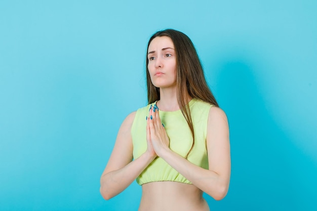 Sad girl is begging by holding hands together on chest on blue background