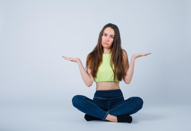 Free photo sad girl is opening wide ehr hands by sitting on floor on white background