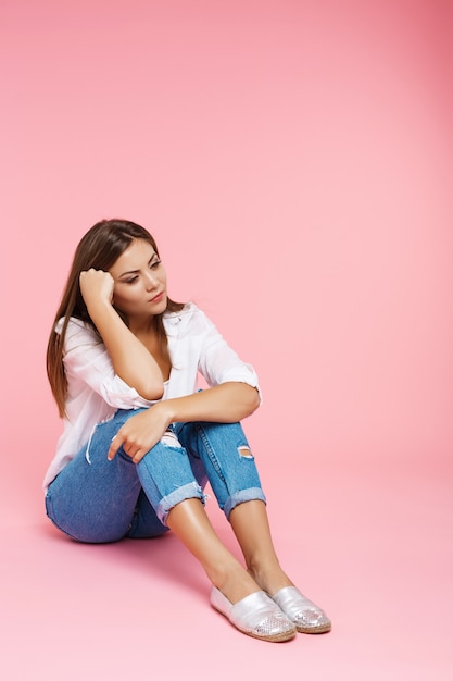 Free photo sad girl sitting on floor looking down isolated on pink