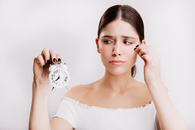 Sad girl in white top looks with sadness at white alarm clock. Portrait of model without makeup.