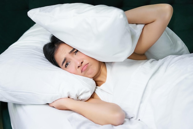 ベッドに横たわっている不眠症の若いアジア女性の悲しい少女は、枕で耳を覆い、パートナーを眠らせない