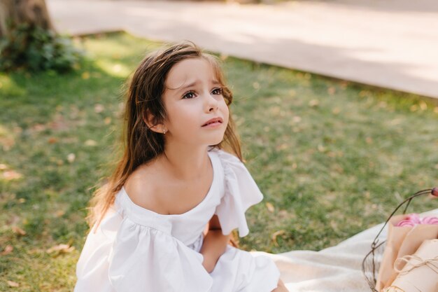 Sad girl with light-brown hair is about to cry sitting on blanket beside alley. Outdoor portrait of unhappy child looking up with eyes full of tears in park.