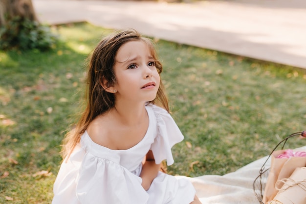 Sad girl with light-brown hair is about to cry sitting on blanket beside alley. Outdoor portrait of unhappy child looking up with eyes full of tears in park.