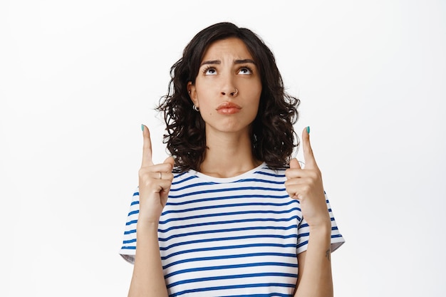 Sad and gloomy brunette woman, frowning upset, pointing fingers up and looking at top advertisement with regret or jealousy, standing disappointed against white background.