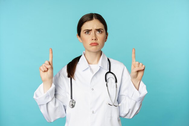 Sad and gloomy doctor, female medical worker sulking, pointing and looking up upset, standing over torquoise background