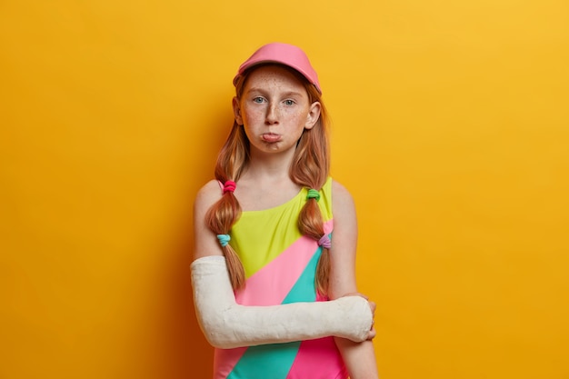 Бесплатное фото Грустная хмурая веснушчатая девочка в красочном купальнике и кепке, сломала руку в гипсе, испортила отдых из-за травмы, изолирована за желтой стеной. летнее время, дети, концепция аварии