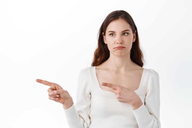 Sad and gloomy girl student sulking grimacing disappointed making upset face and pointing fingers aside at copyspace for your banner logo standing against white background