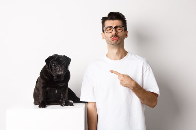 Sad and gloomy pet owner pointing at his black pug dog and sobbing, standing upster against white background
