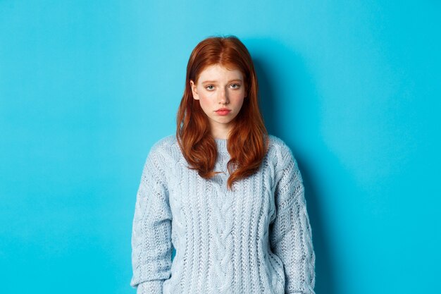 カメラを不安に見つめ、気分が悪く、セーターの青い背景に立っている、悲しくて暗い赤毛の10代の少女。