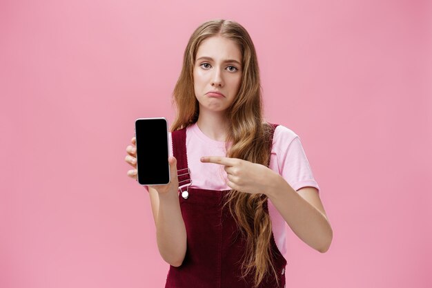 人差し指でガジェットを指さしているスマートフォンの画面を示すかわいい波状の自然な髪型の悲しい憂鬱な若い女性は、壊れた電話を購入した後、不機嫌な顔をしかめている。