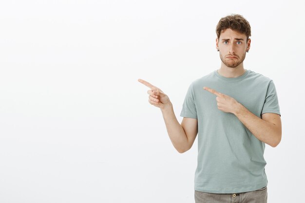 Sad gloomy young man in t-shirt, making upset disappointed face while pointing left with index fingers, frowning, being unhappy and miserable