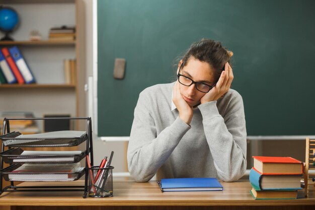 sad grabbed head young male teacher sitting at desk with school tools on in classroom