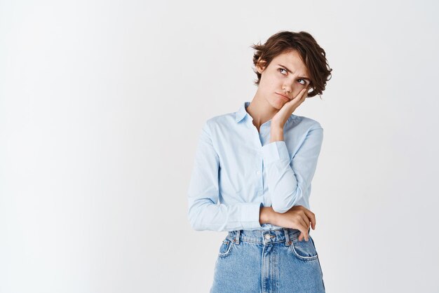 Sad and grumpy young woman in shirt looking at upper left corner with offended angry face frowning and standing jealous on white background