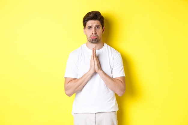 Sad guy begging for something, sulking and asking for favour, saying sorry, standing over yellow background.