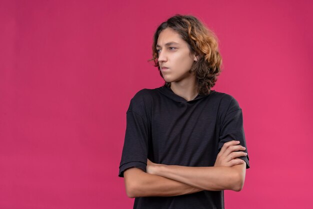 Sad guy with long hair in black t-shirt crossing his hand on pink wall