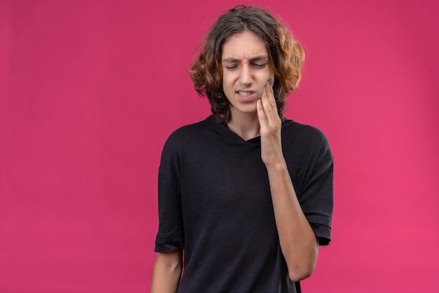 Sad guy with long hair in black t-shirt put his hand on aching tooth on pink wall