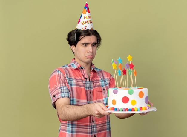 Un bell'uomo caucasico triste che indossa un berretto da compleanno tiene e guarda la torta di compleanno