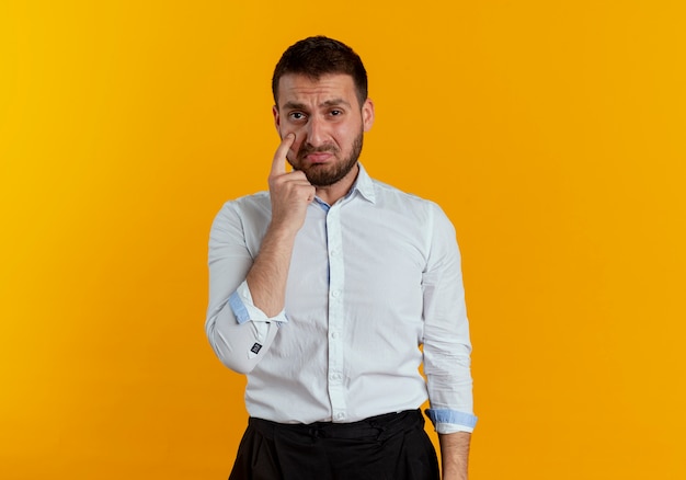 Sad handsome man points at eye isolated on orange wall