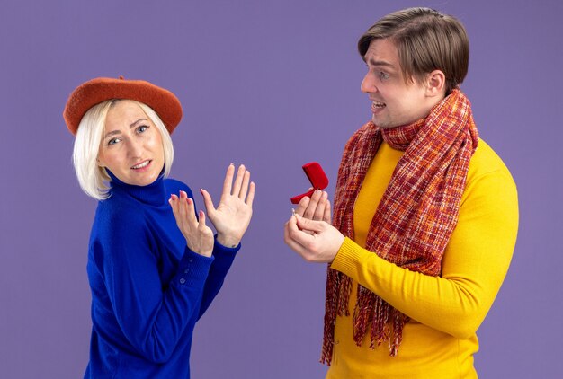 Sad handsome slavic man with scarf around his neck holding red ring box and looking at unpleased pretty blonde woman with beret on valentine's day 