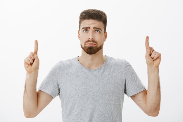 Free photo sad handsome and timid cute masculine adult man with beard and blue eyes looking and pointing up frowning feeling gloomy staring at unfair thing, standing envious and displeased over gray wall