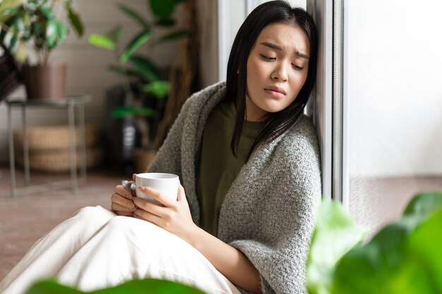 熱いお茶を飲み、悲しんでいる窓の近くの床に座っている悲しい失恋したアジアの女性