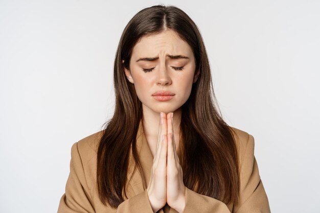 Грустная обнадеживающая деловая женщина молится, закрывает глаза и умоляет, закрывает глаза и умоляет, стоя на белом фоне