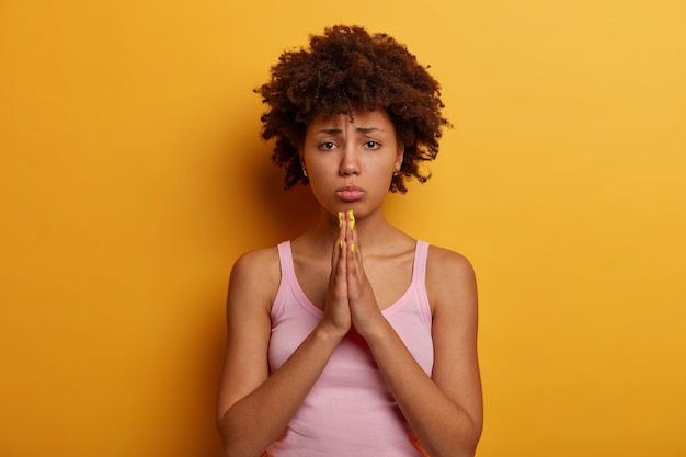 Sad hopeful woman beggs for your help, presses palms together in pray gesture, says please and asks to make favour, purses lips, dressed in casual outfit, feels need, isolated on yellow wall