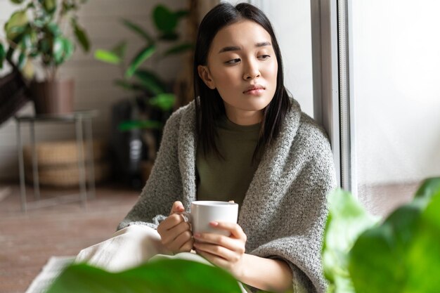 Sad ill asian girl staying on self quarantine during covid catching flu and sitting at home with cup...