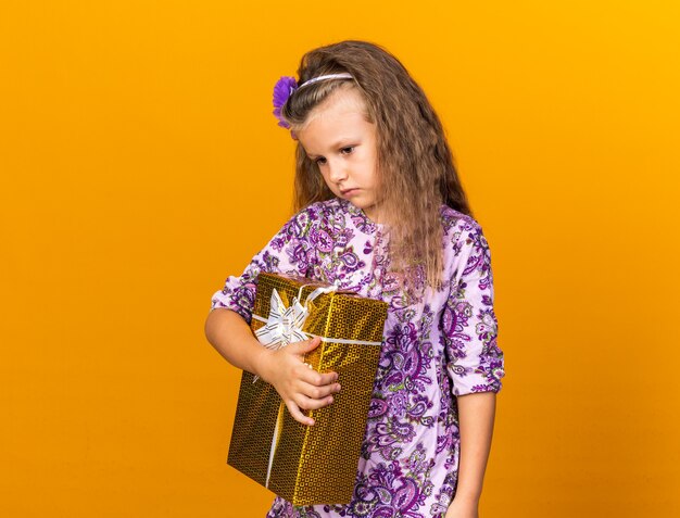 sad little blonde girl holding gift box isolated on orange wall with copy space