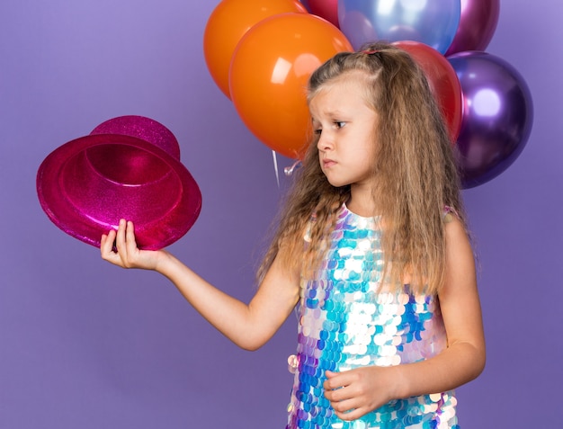 sad little blonde girl holding and looking at violet party hat standing with helium balloons isolated on purple wall with copy space