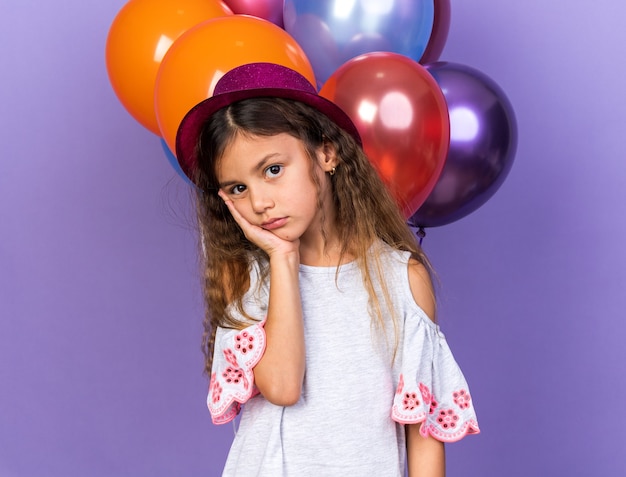 sad little caucasian girl with violet party hat putting hand on face standing in front of helium balloons isolated on purple wall with copy space