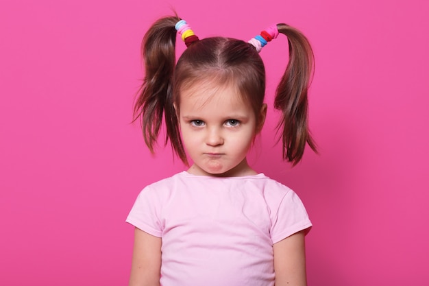 Sad little girl stands on pink wall. cute kid wears rose t shirt, has two fanny poni tails with many colourful scrunchies, looks hurt with pouty lips. upset child on playground.
