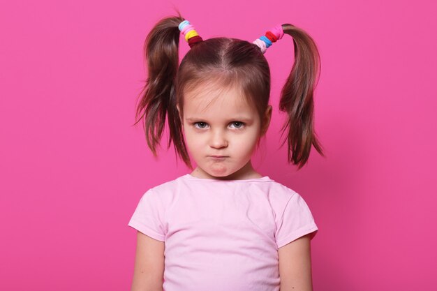 Sad little girl stands on pink wall. Cute kid wears rose t shirt, has two fanny poni tails with many colourful scrunchies, looks hurt with pouty lips. Upset child on playground.