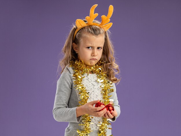 Sad little girl wearing christmas hair hoop with garland on neck holding christmas balls isolated on blue background